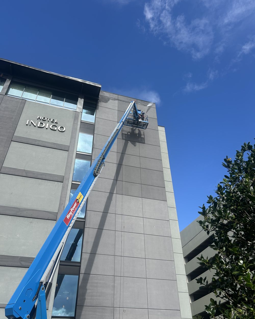 Soft washing the upper floors of a multi-storey commercial building from an extended boom lift, spray visible against a clear blue sky.