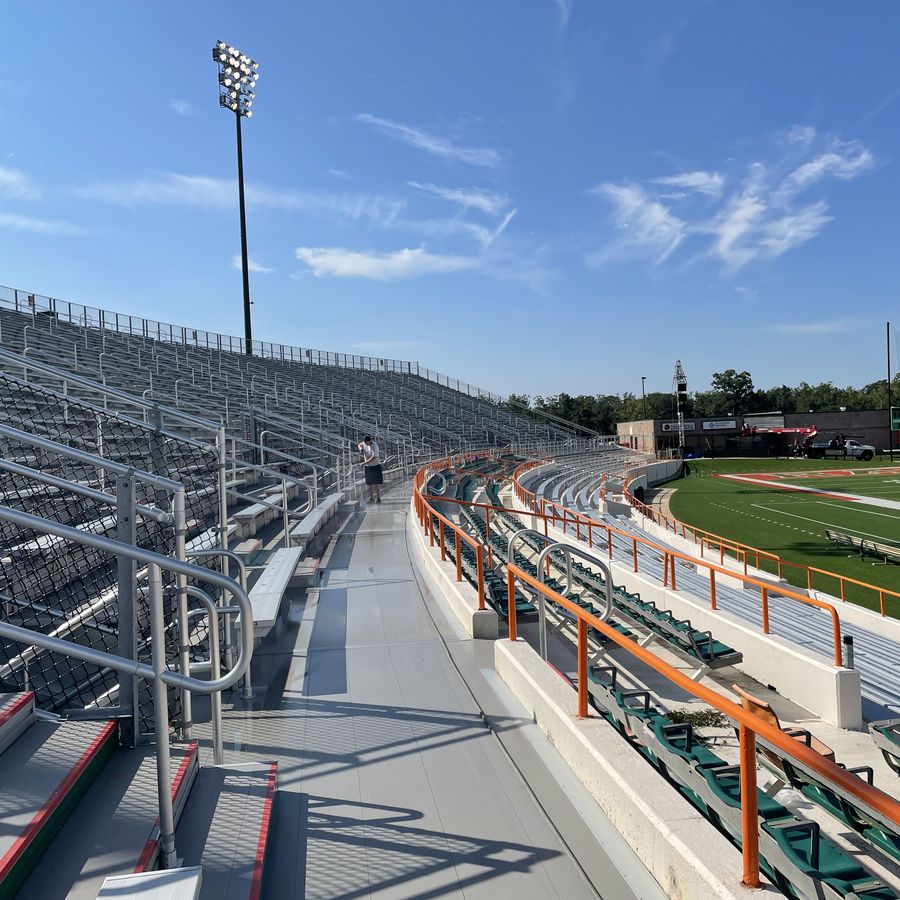 Clean aluminium stadium bleachers and painted handrails curving alongside a sports field.